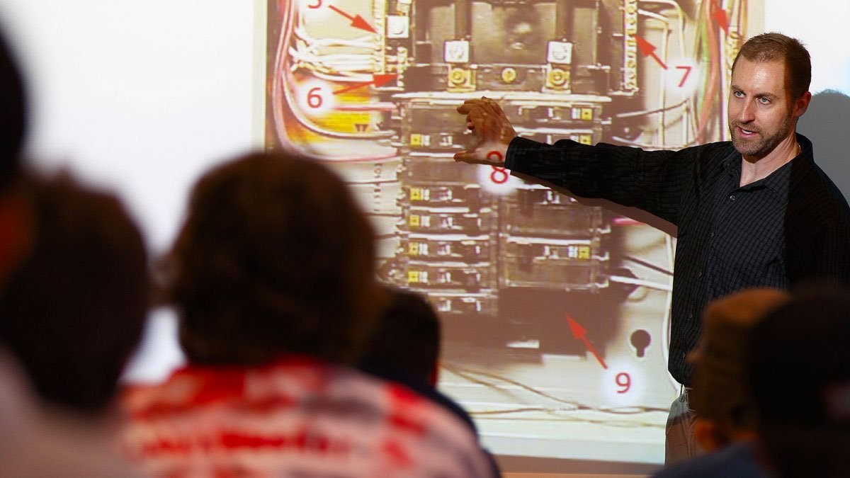 Male professor stands in front of a class pointing at at image on the white board