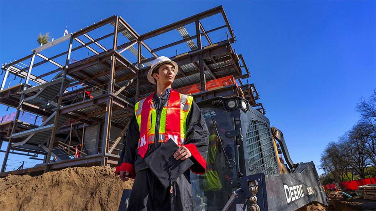 Student in hard hat and construction vest stands in front of an unfinished bridge.