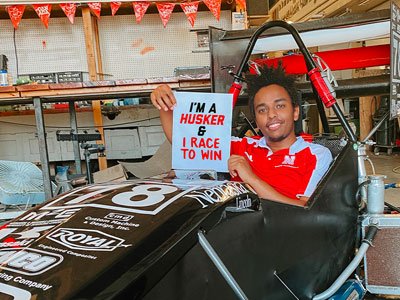 Male student sitting in a SAE car holding a sign: "I'm a Husker & I Race to Win"