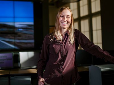 Sydney stands next to a computer and large screen showing a highway